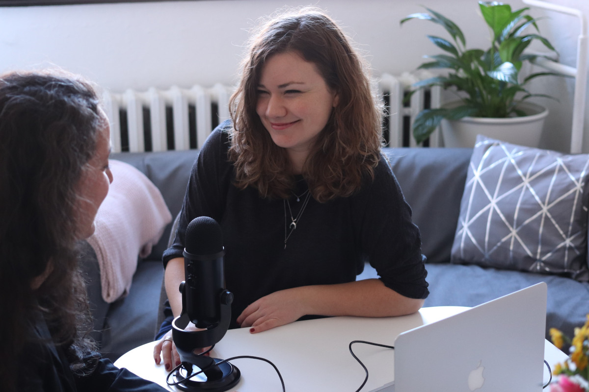 A woman in black half-sleeved shirt sitting while facing woman and smiling. They are stilling at a desk which has a black USB microphone and an Apple laptop.