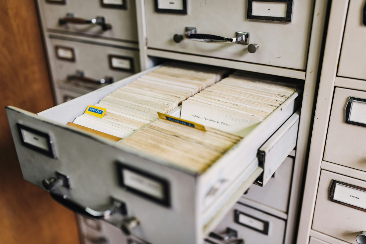 In the image, you can see an open file cabinet with numerous drawers. The top drawer appears to be filled with files, and there's a white label or tag attached to it, though the text on the label is not fully visible. The cabinet itself has a beige finish and features metal handles for each drawer. The drawers are arranged in three vertical columns, and the cabinet occupies most of the frame from left to right. The setting suggests an office environment where records or documents are stored.