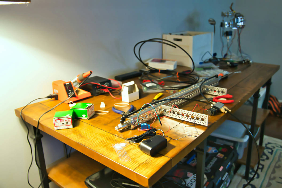 This image depicts a workspace that appears to be focused on electronics or hobbyist projects. There is a wooden table in the foreground with various items scattered across its surface, including what seem to be electronic components, tools like pliers and wires, and some small electrical devices. On the left side of the table, there's a white box that may contain additional components or supplies. The right side of the image shows a desk with similar items, suggesting that this is a common area for work in this space. In the background, there's a glimpse of a room with a bed and other personal belongings, indicating that this might be a private residence where electronics hobbies are pursued. The setting appears to be casual and lived-in, reflecting a personal workspace rather than an organized or professional environment.