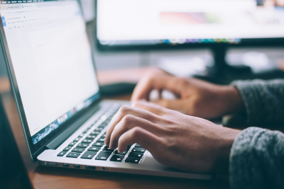 The image is a photograph depicting a blurred person working on a laptop computer. The individual appears to be typing or using the keyboard and touchpad, indicated by their hand hovering over these input devices. The laptop screen is partially visible but mostly obscured, making it difficult to discern any content displayed there.
The workspace has a clean and minimalist aesthetic, with a wooden desk providing support for the laptop. There are no other items on the desk except for a desktop computer monitor in the background on the right side of the image. The monitor is off, showing a dark screen.
The room has neutral colors, suggesting a professional or modern office environment. Natural light appears to be entering the space from the left side, indicating it could be a window or an open door out of frame.
In the foreground, there are two keyboards visible; one on the laptop and another that seems to belong to a keyboard-less desktop computer, which is not in focus. The focus and depth of field are shallow, causing parts of the image to appear blurred, which draws attention to the person's hand on the laptop keyboard as a focal point.