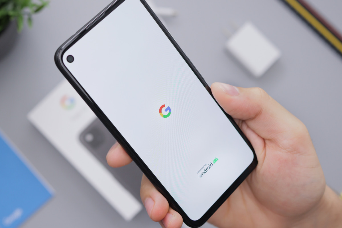 A mobile phone showing a white screen with the Google logo in the centre. The phone is held by a caucasian hand, and in the background (out of focus) is a grey table with the box for the phone, a potted plant, and the phone charger.