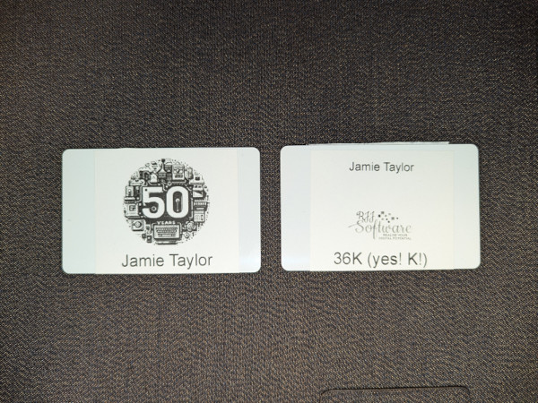 The image shows two name badges placed next to each other on what appears to be a table.
On the right, there's a badge with the name 'JAMIE TAYLOR' prominently displayed at the top center, and below that is an additional line of text with '36K! (yes! K!)', and between them is the RJJ Software logo.
The badge on the left has the words '50 years' at the top center, as part of the logo for the event. Below this is the name 'JAMIE TAYLOR' again, indicating that both badges are for the same person.
Both badges have a rectangular shape and appear to be made of plastic.