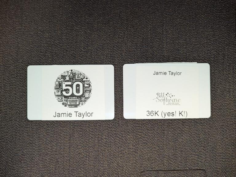 The image shows two name badges placed next to each other on what appears to be a table.
On the right, there's a badge with the name 'JAMIE TAYLOR' prominently displayed at the top center, and below that is an additional line of text with '36K! (yes! K!)', and between them is the RJJ Software logo.
The badge on the left has the words '50 years' at the top center, as part of the logo for the event. Below this is the name 'JAMIE TAYLOR' again, indicating that both badges are for the same person.
Both badges have a rectangular shape and appear to be made of plastic.