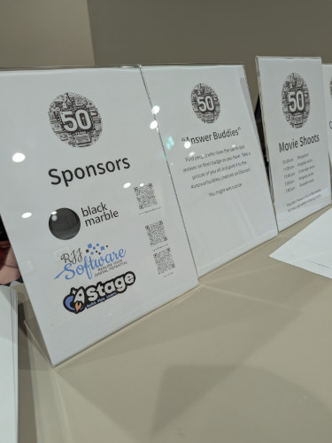 The image shows a display featuring posters of sponsors or exhibitors at an event. At the top, there is a large white poster with black text that reads '50 Sponsors' and below it, the names of the companies are listed in black text on a white background. Each company name is accompanied by their logo or symbol. To the right, there's another smaller white poster displaying the logos of the sponsors. The overall style of the image is informational and appears to be taken at an event where such displays are common.