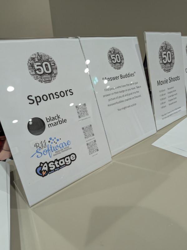 The image shows a display featuring posters of sponsors or exhibitors at an event. At the top, there is a large white poster with black text that reads '50 Sponsors' and below it, the names of the companies are listed in black text on a white background. Each company name is accompanied by their logo or symbol. To the right, there's another smaller white poster displaying the logos of the sponsors. The overall style of the image is informational and appears to be taken at an event where such displays are common.