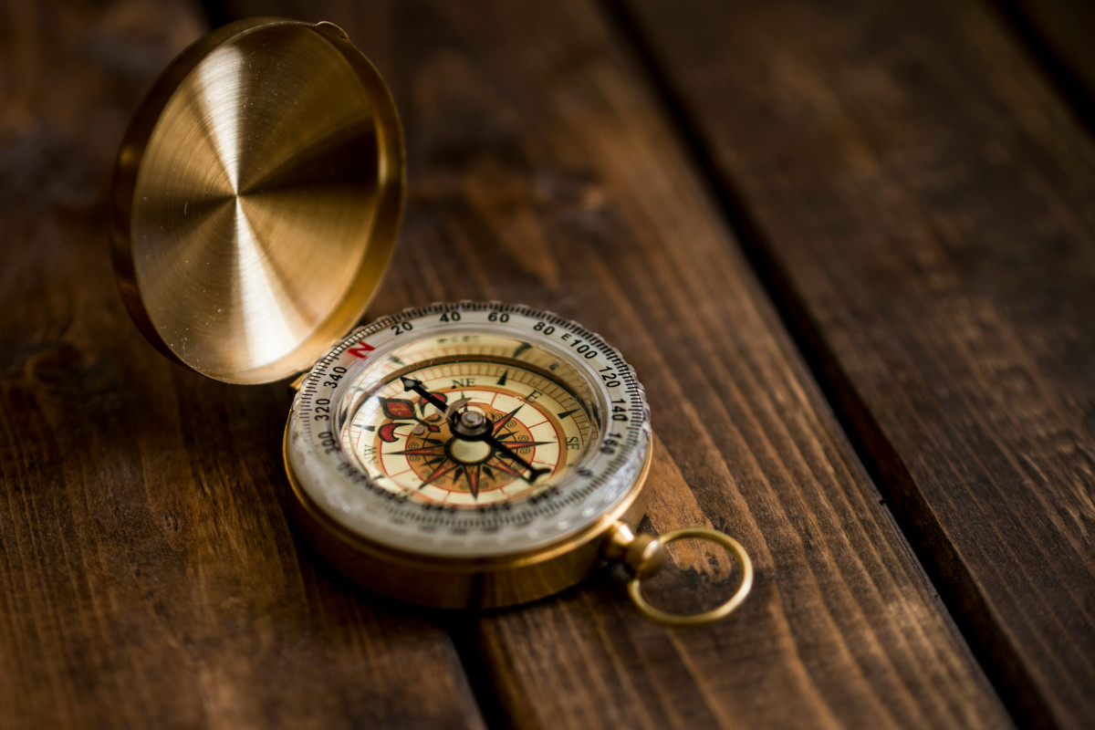 The image presents a compass, a symbol of guidance and navigation. The compass has a gold casing with a silver needle, indicating its precision and durability. It's set against a rustic wooden background, which adds to the vintage charm of the object. The needle of the compass is pointing towards the top left corner of the image, suggesting an unseen destination or goal.