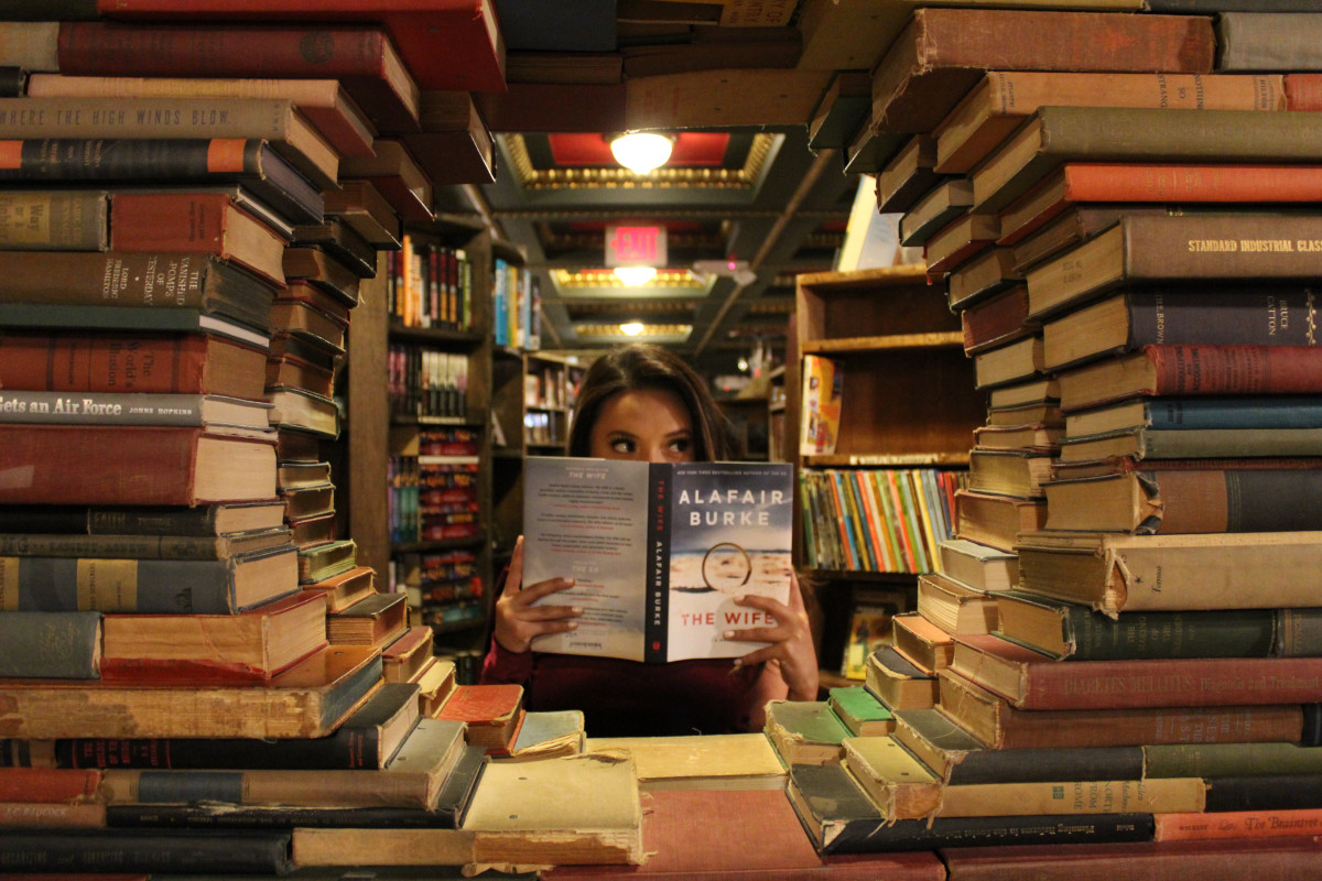 A woman holding a book, pretending to read it, whilst looking off to the right of the image. The woman is sitting in a library and is surrounded by books. She is framed by a number fo books arranged to provide a circular window.