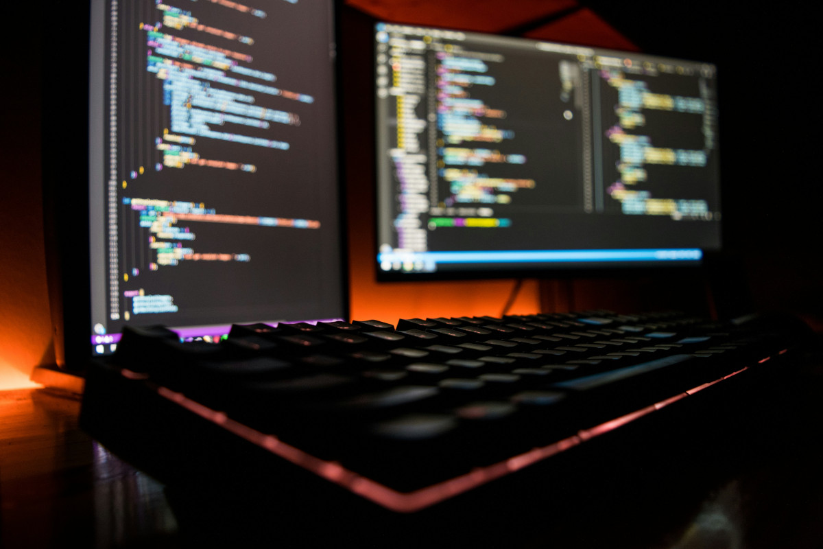 The image shows a desktop computer setup with two computer monitors displaying code in a terminal window. The monitors are  placed side by side on a desk, and between them is a keyboard with a pink backlight. There is on keyboard visible; partially shown in the foreground. On the left side, there's a wall with a red light emanating from it, which appears to be glowing on the desk and under the keyboard as well. The image has an overlay effect where two photos are superimposed, creating a dual exposure appearance.