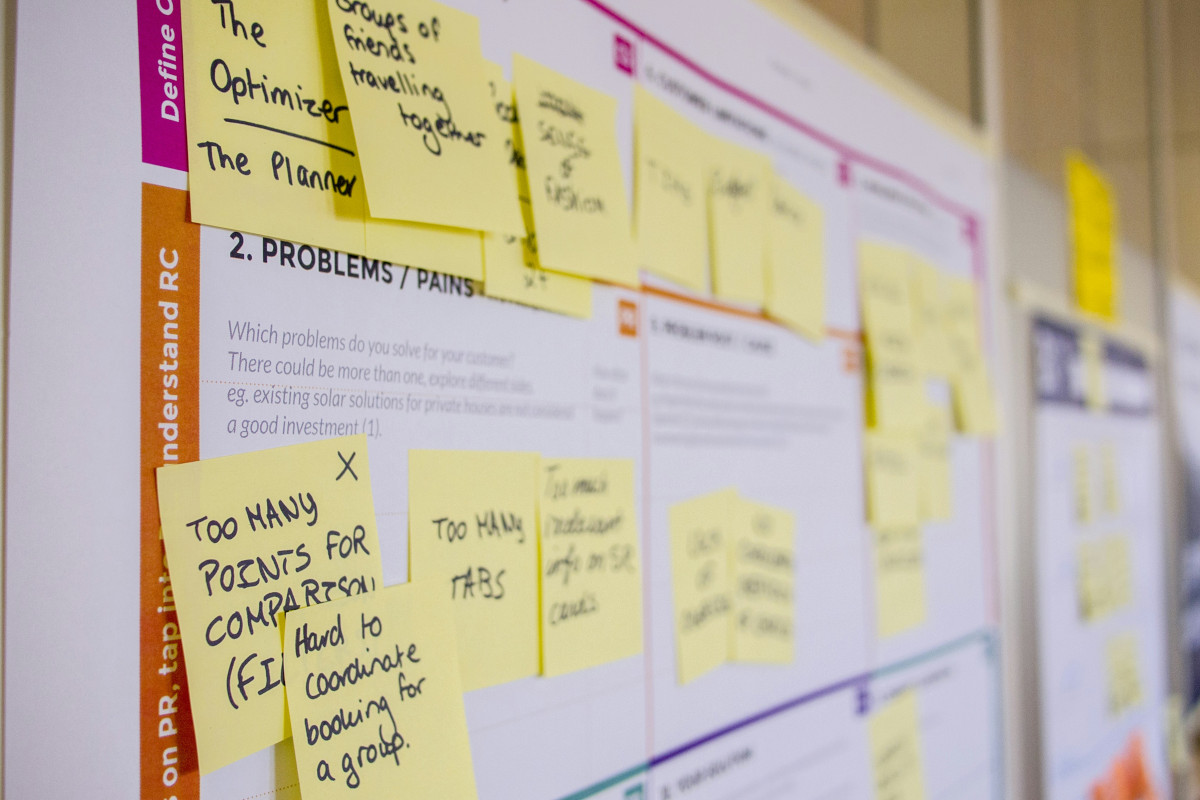 The image shows a wall covered with multiple sticky notes, each containing different handwritten text. Some of the visible texts include 'THE SOLUTION TO THE PROBLEM', 'NEED TO COMPARE BOOKING TO...', 'CONCLUSIONS FOR A FULL COMPARISON', and 'TOO MANY TABS'. The sticky notes are grouped together, suggesting an ongoing brainstorming session or planning activity. The wall is set against a background that appears to be a whiteboard or board with some writing and diagrams on it, further indicating a workspace or project area. The image has a candid quality, as if capturing an unplanned moment of work in progress