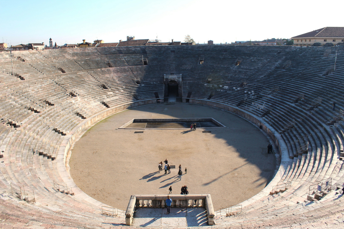 The image depicts an ancient amphitheater under the sun. It's a large, circular structure with multiple levels of seating arranged around a central stage or pit. The audience areas are made up of rows of stone seats, and the walls surrounding the arena show signs of weathering, indicating its historical significance. In the foreground, there is a crowd of people standing in front of the amphitheater, possibly spectators or tourists. Some individuals appear to be engaged in various activities, such as taking photographs or walking around.