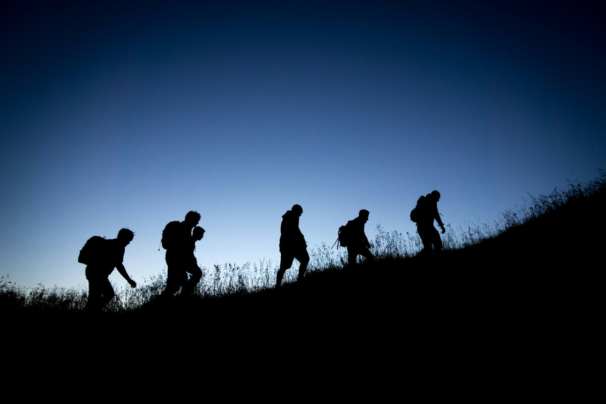 The image depicts a serene landscape with lush green trees and foliage filling the scene. In the foreground, in silhouette are six people on a hike, they are hiking uphill. We cannot make out many details about them, other than there are six of them. In the background, there is a clear blue sky that extends to the horizon. A few clouds are scattered across the sky, adding depth and texture to the image. The overall composition of the image suggests a peaceful and natural setting.