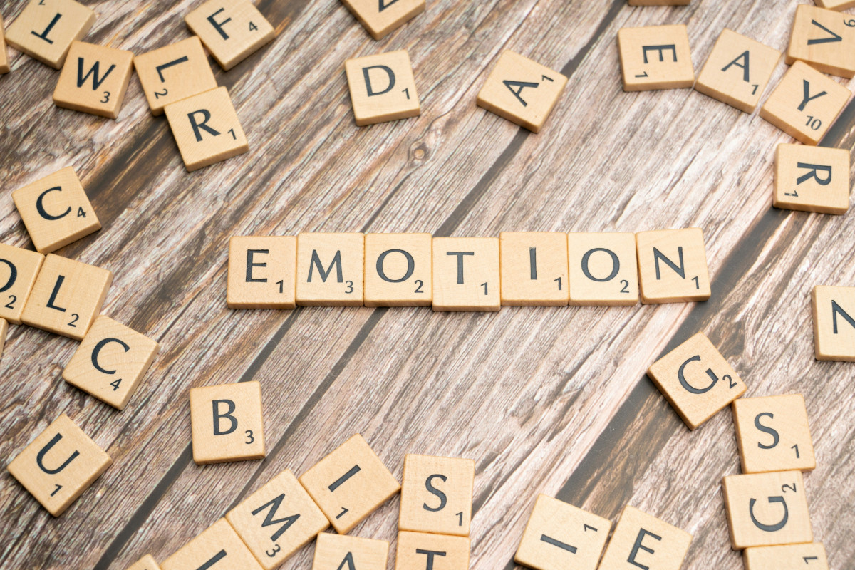 This is a photo of an assortment of Scrabble tiles spelling out the word 'emotion' in the center. The tiles are placed on a surface that appears to be wooden, creating a circular arrangement around the central word. The background is blurred and not distinctly clear, but it seems to suggest a table or platform setting. The overall image conveys a theme of vocabulary, language, or perhaps emotional expression as indicated by the word 'emotion.'