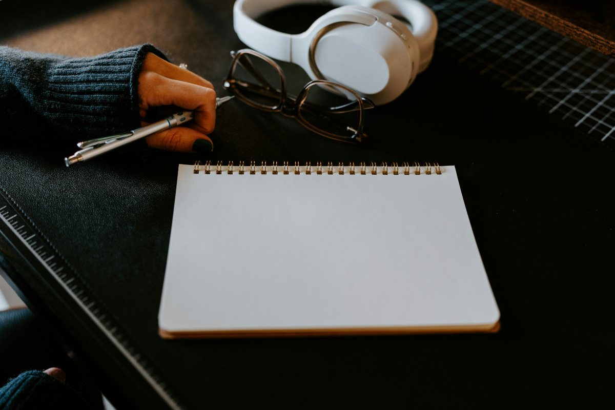The image shows a close-up of a person's hands working on a piece of paper. The paper appears to be a blank notepad with yellow-golden lines indicating its grid structure. On the desk where this activity is taking place, there are several items including a pair of glasses, a pen, and what looks like an eye patch or perhaps some kind of medical equipment. writing, planning, or note-taking.