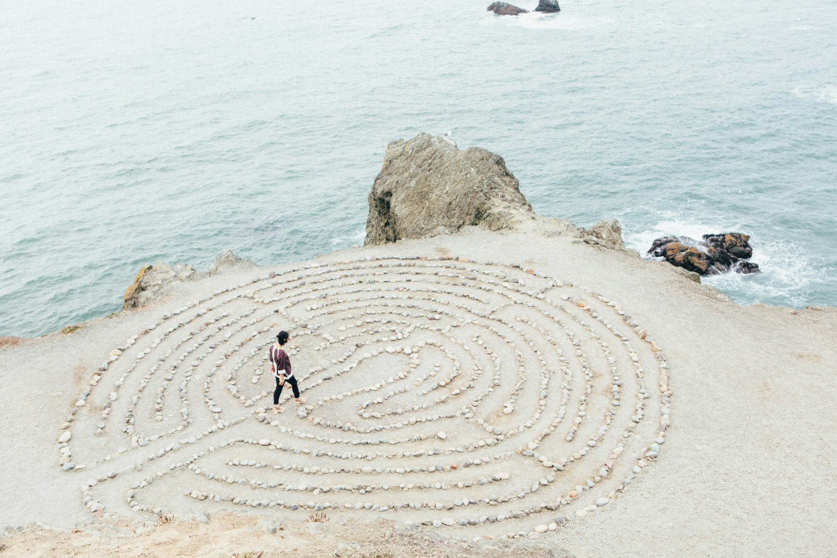 The image depicts a tranquil coastal scene. In the foreground, there's a large stone labyrinth situated on a cliffside overlooking the ocean. The labyrinth has concentric circular patterns that are intricate and detailed. A person stands at the entrance of the labyrinth, looking out towards the water, giving a sense of scale to the size of the structure. Beyond the labyrinth, the coastline is visible with waves crashing onto the shore, and in the distance, there's a rocky outcropping extending into the ocean. The sky above is overcast, with no visible clouds, suggesting a calm day. There are no visible texts or distinctive brands in the image that would require a detailed description.
