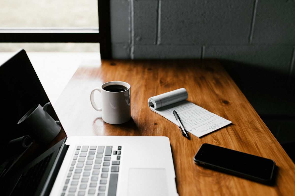 In the image, there's a desk set up in what appears to be a cozy room. The desk is made of wood and holds two items: a cup of coffee, which suggests someone might be working or studying here, and a notepad with a pen resting on it, indicating that writing or note-taking is being done. There's also a laptop on the right side of the table, hinting at a blend of traditional and modern work methods. The room has a warm ambiance, with a window in the background letting in natural light. The desk is positioned against a wall, creating a sense of space within the room. The overall setting implies a quiet, comfortable environment conducive to focus and productivity.