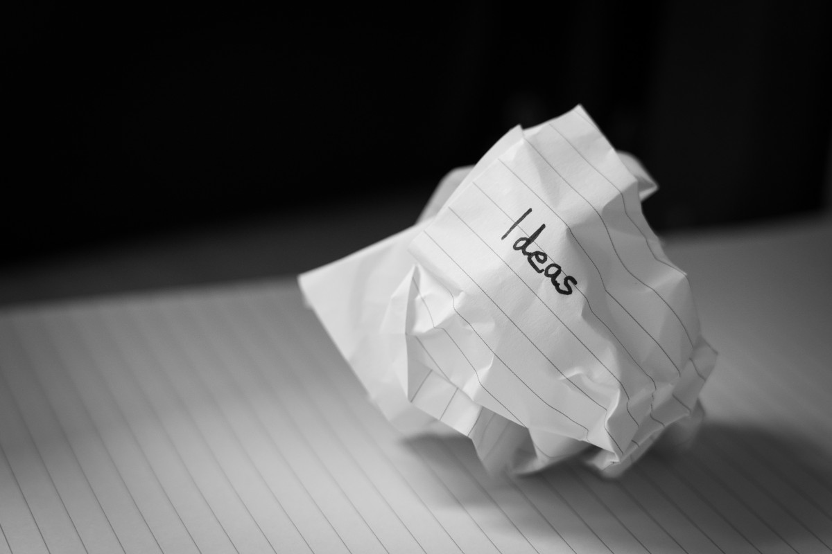 The image shows a paper crumpled into a ball and resting on a surface, possibly a table or desk, with visible notepad lines in the background. On top of the paper is handwritten text that reads 'ideas' in a somewhat messy handwriting style. The ball of paper appears to be placed there deliberately, as if it's been crumpled and tossed aside after a brainstorming session or idea gathering activity. There's also a faint reflection of light on the surface beneath the paper. The lighting suggests an indoor setting with ambient artificial light.