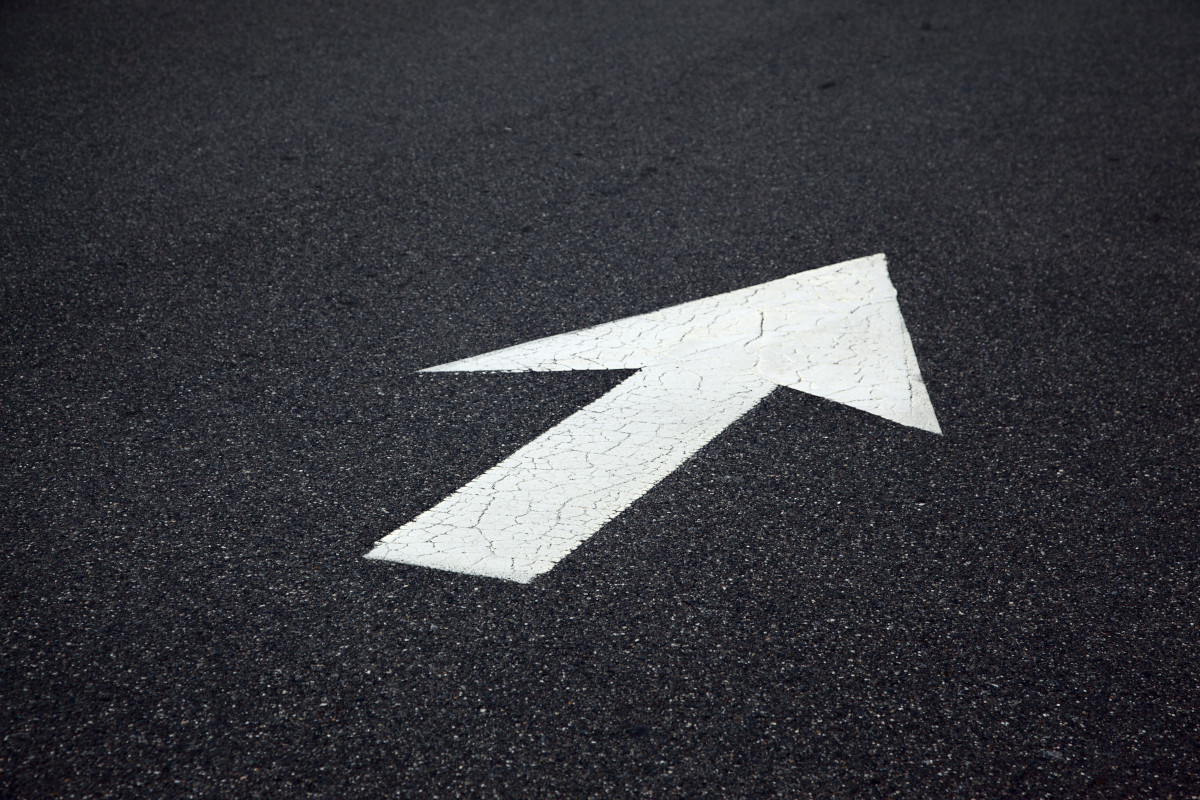 The image shows a stylized road sign arrow against a backdrop that appears to be an asphalt surface with some texture suggesting it might be tarmac. The arrow is white and pointed upwards, indicating the direction of traffic or movement in  that direction. The sign has a black outline and there are no visible texts on it. The style of the image is graphic, with a focus on the simplicity and clarity of the sign itself. 