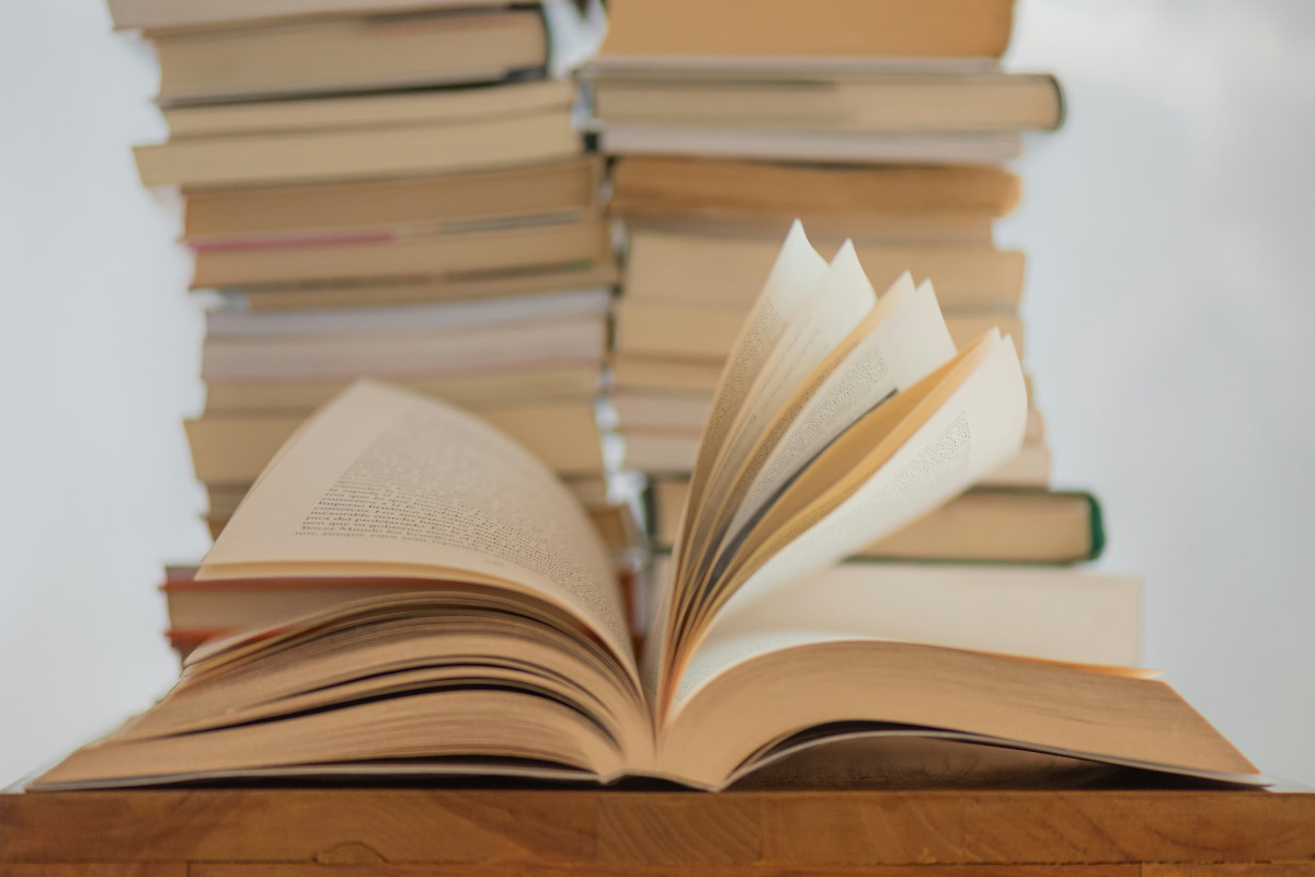 A collection of books stacked on top of each other in an indoor setting. The books come in various sizes and colors, indicating a diverse range of titles or genres. The photo has a vertical orientation with the spine of a book facing outward prominently.
