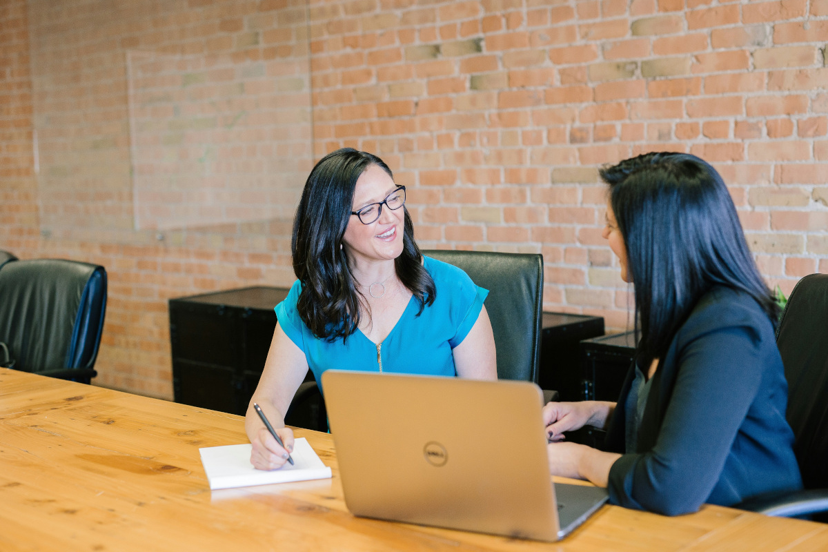 Two women sitting at a desk in an office environment. The woman on the left is smiling and leaning forward slightly, while holding a pen and appears to be writing or about to write something. She is wearing a blue top, has dark hair, and is dressed casually. The woman on the right is attentively listening, wearing glasses, and has her hand resting on the desk. There's an open laptop in front of her, suggesting she may be involved in some work or project discussion. Both women are positioned in front of a brick wall with a mirror reflecting part of the room. The space looks professional yet welcoming, with a cozy ambiance.