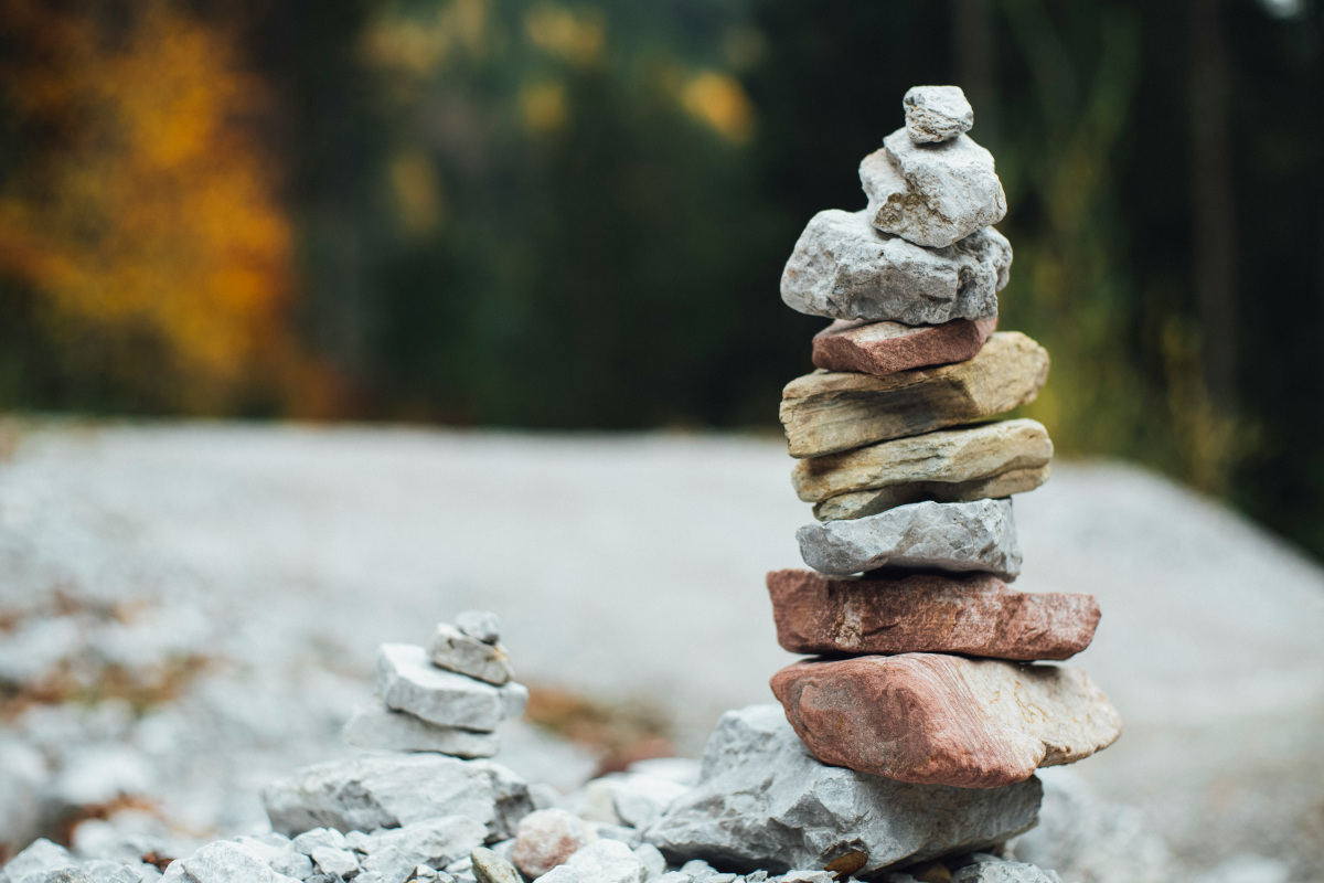 This is an image of a rock stack composed of variously shaped and sized rocks. The rocks are balanced precariously atop one another in what appears to be an outdoor setting, possibly near a natural environment with vegetation and rocks in the background. The central focus of the image is on the stacked rocks themselves, which display a range of colors from earthy browns and yellows to greys and white. The rocks are arranged in an irregular pattern with some stacked vertically and others horizontally, creating an unstable yet harmonious balance. There's no text or other discernable objects within the image that would provide additional context or information. 