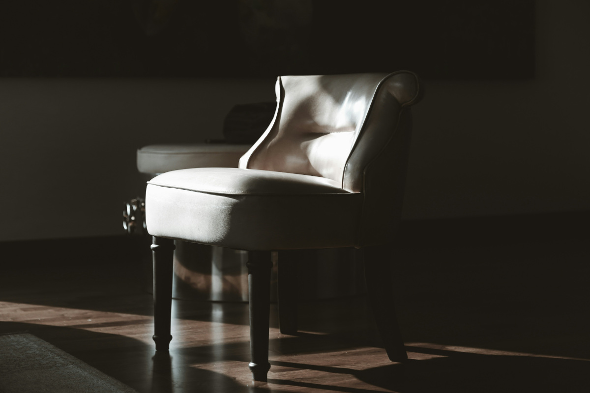 The image features a wooden chair placed in front of a large window. The sunlight filters through the glass panes, casting a warm glow onto the chair and creating a dramatic contrast between the illuminated areas and the shadows. The chair is styled with a curved backrest and armrests, suggesting a sense of comfort and elegance.