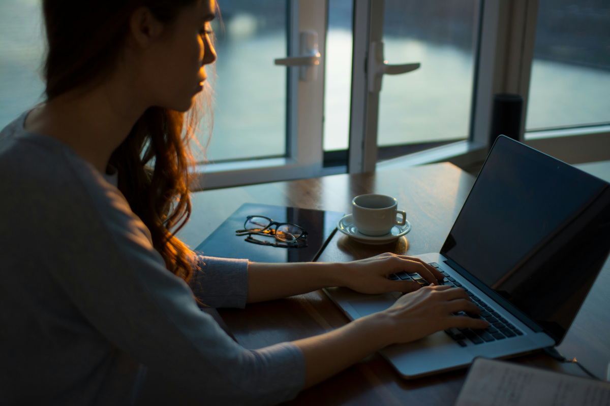 The image depicts a woman sitting at a desk in an indoor setting. She is working on a laptop that is open and placed centrally on the desk. In front of her, there's a coffee cup suggesting she might be having a drink while working. There are two windows visible: one near the coffee cup and another to the left. The windows appear to be closed. The room has modern decor; there is a framed picture on the wall above the desk, a plant to the right of the desk, and a wooden side table to the left. Natural light seems to be coming through one window, illuminating part of the room. The overall mood of the image is serene and focused, with the subject engaged in work or study. There is no visible text within the image.