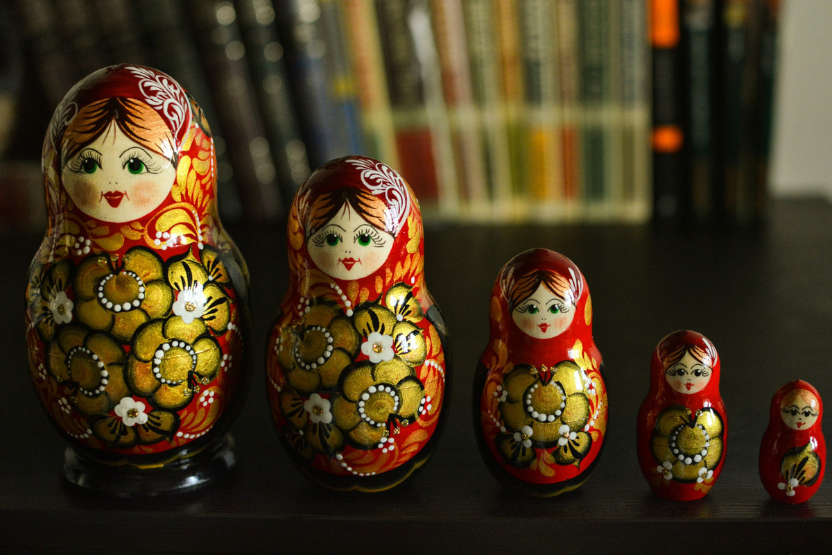 The image shows a charming set of matryoshka dolls, which are traditionally Russian nesting dolls. There are three dolls in the foreground and one larger doll in the background. Each doll is painted with vibrant colors and patterns, and they all feature traditional clothing, suggesting a cultural theme. They seem to be placed on a flat surface, possibly a table or shelf, and in front of them, there appears to be a book with a cover that features what looks like a character resembling the dolls. The background is blurred but suggests an indoor setting, possibly a room with some bookshelves or a similar display.