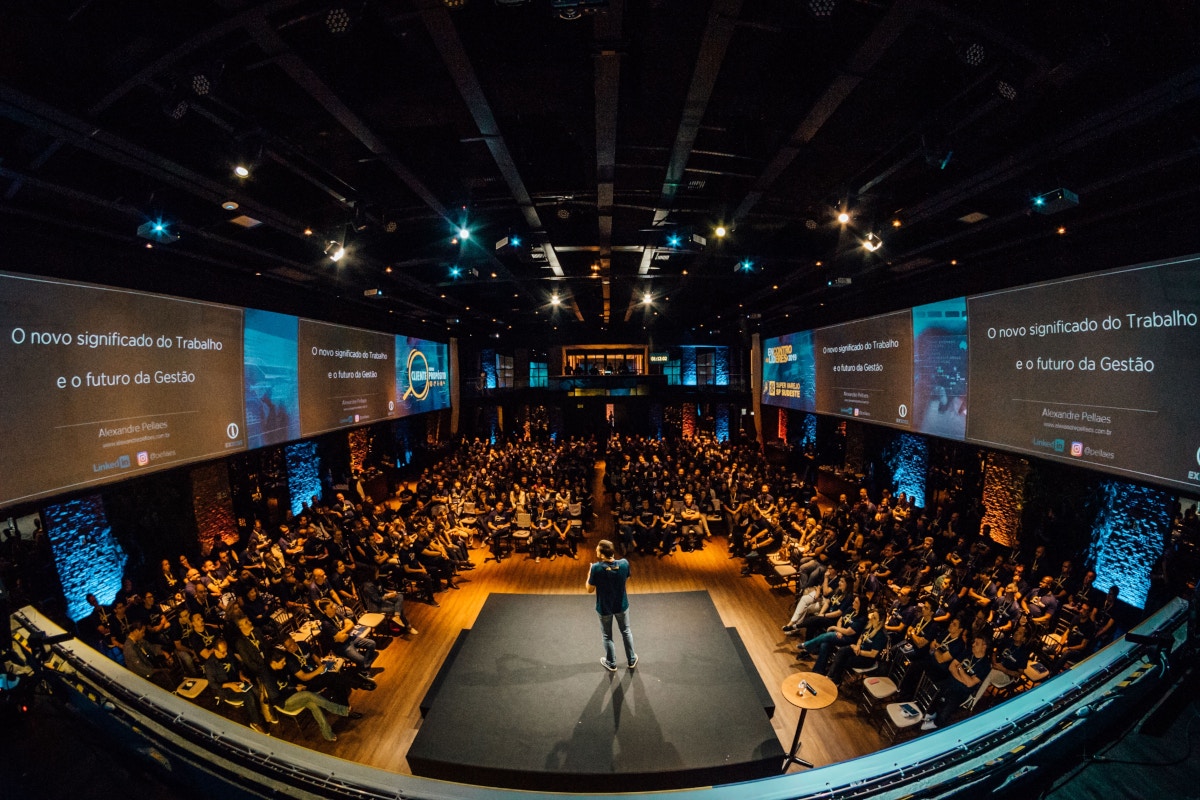 A panoramic photo showing the attendees of a talk watching the speaker. The speaker is standing on a raised platform and looking to their left. The camera is placed behind and above the speaker, looking down on the audience, and there are screens above the audience on the left and right hand sides of the speaking space. There is also a hint of a screen behind the speaker - shown at the very bottom edge of the frame.