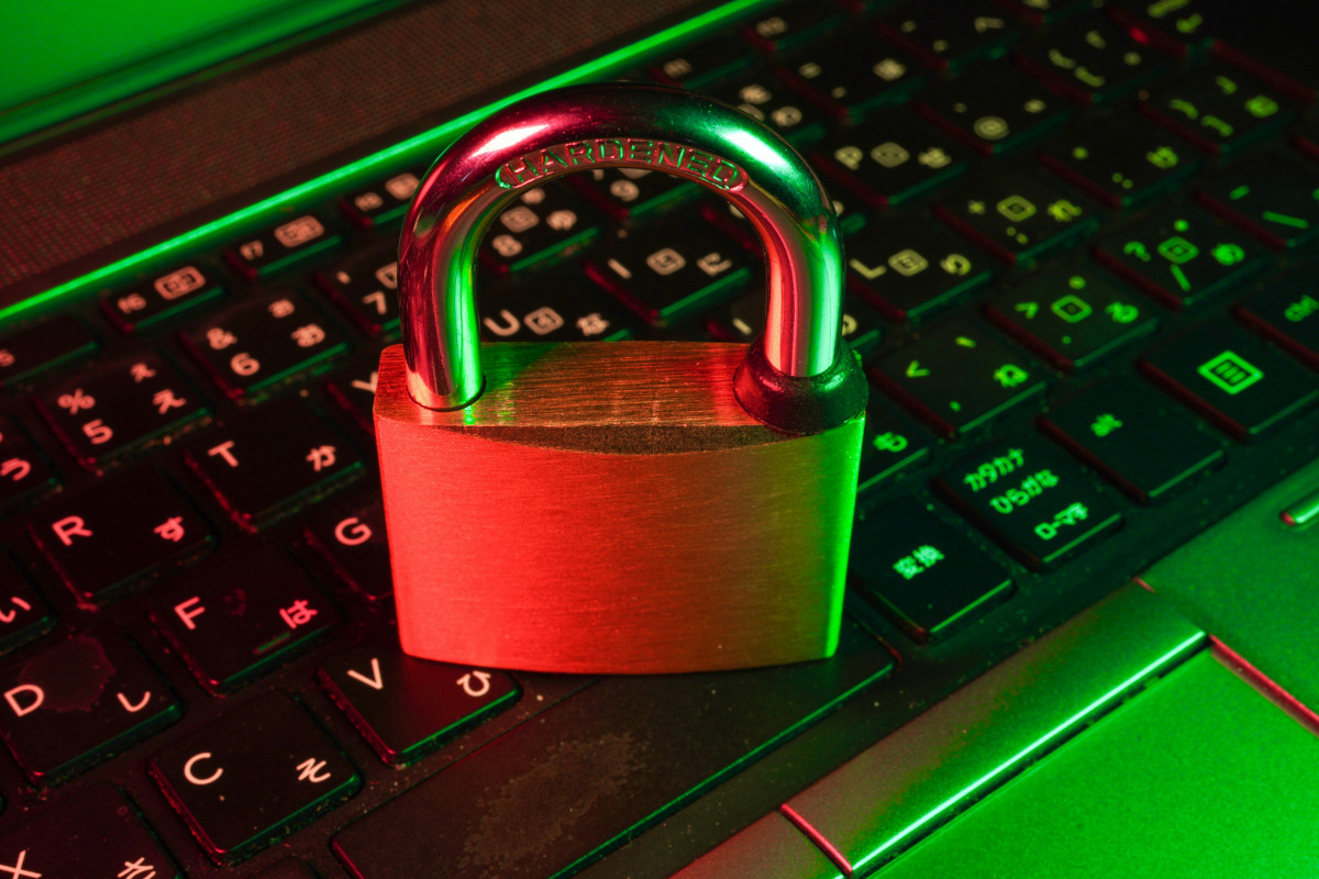 A brass padlock with the word 'HARDENED' stamped on its steel shackle, resting on a laptop keyboard with Japanese and English key legends. The scene is lit in contrasting red and green light, with the red falling on the padlock's body and the green glowing across the keys.