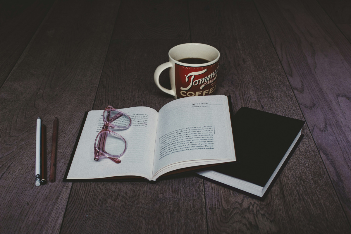 A black and white photograph showcasing an open book with pages facing outward, resting on top of two opened notebooks. Beside them is a mug of coffee, placed next to a red pen and glasses, suggesting someone has recently been reading or studying in this space. The background features a light-colored hardwood floor and a portion of a wooden table, creating an ambiance conducive to focused work or study.