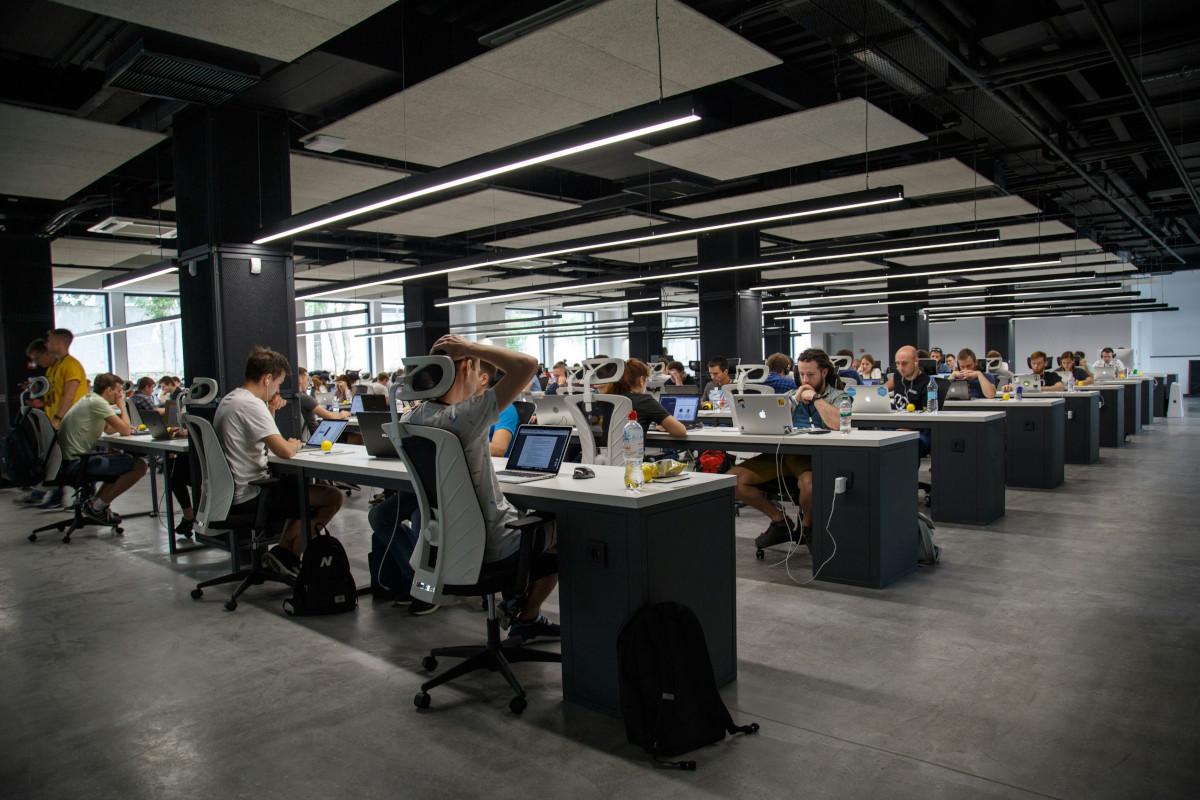 This is a photograph of an office interior with a contemporary design. The space features rows of desks, each equipped with a computer monitor, a keyboard, and a chair. A few individuals are seated at the desks, working or using their laptops. In the background, there is a large window allowing natural light to enter the room, which has an industrial feel due to its exposed ceiling and concrete flooring. The overall atmosphere suggests a modern workspace designed for collaboration and productivity.
