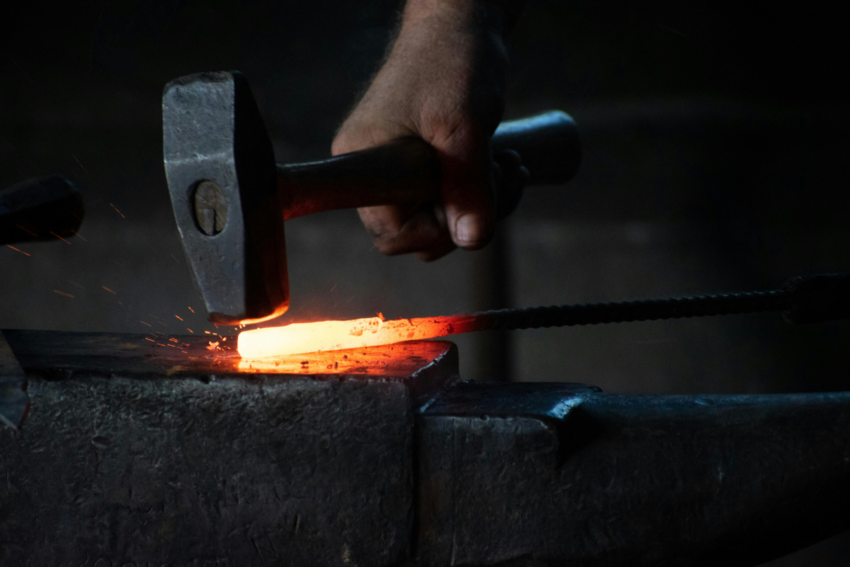 In the image, there is a blacksmith at work. The blacksmith is holding a large hammer in one hand and a piece of red-hot metal in the other. The blacksmith's tools are on anvil, which has been heated to create a glowing ember effect. Sparks can be seen flying from the metal as it is being worked upon, emphasizing the intensity of the process. The setting appears to be a traditional forge with a dark, moody ambiance typical of a blacksmith's shop.