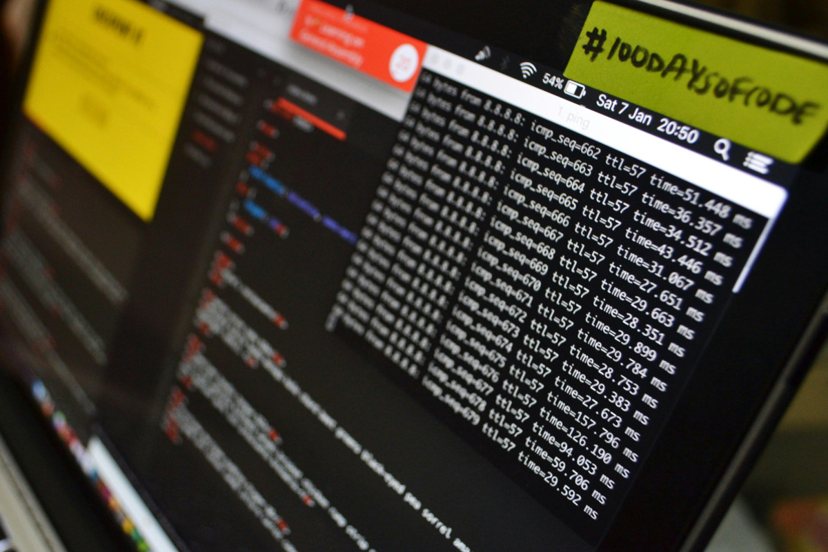 The image displays a computer monitor that is displaying multiple browser tabs open. Each tab appears to show a webpage with a table or list of items, possibly related to software development or IT management. Below these open tabs, there's a sticky note attached to the  monitor that reads '#100DaysOfCode.' The computer screen is illuminated and takes up most of the image, suggesting the focus on the digital content displayed. The lighting in the room seems soft and ambient, casting gentle shadows under the monitor and on the desk it sits upon. In the lower right corner of the image, there's a glimpse of a smartphone lying on its side. The overall atmosphere suggests a workspace where someone is actively working on coding projects, with a particular focus on CodeDays events or challenges. The sticky note indicates that this space might be used for self-improvement or community-driven code learning activities.