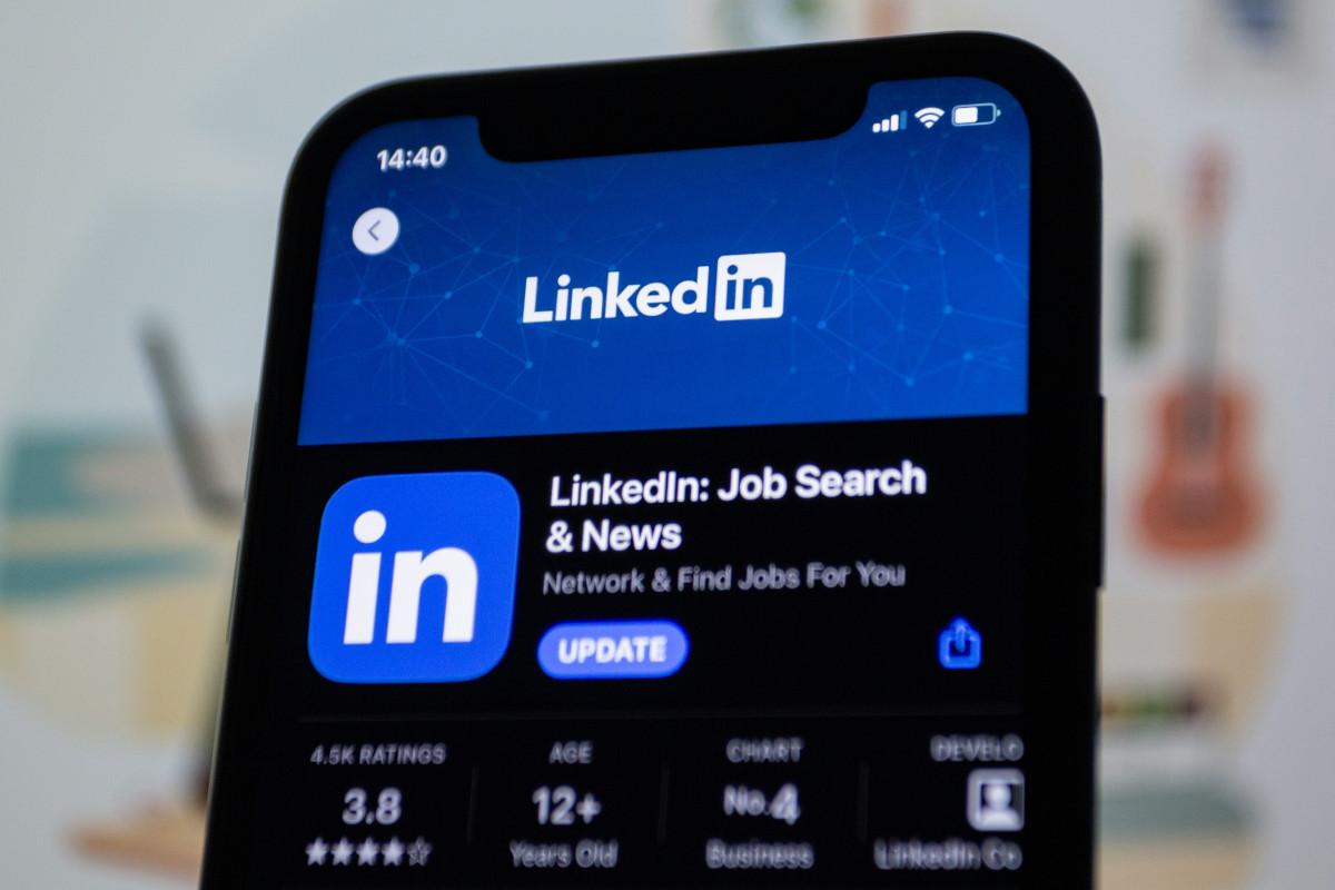 The image shows a smartphone displaying the LinkedIn app interface. The main part of the screen is showing a search result with the LinkedIn logo and the text &quot;LinkedIn News & Jobs&quot; beneath it. There are other elements visible in the background, including another phone screen to the left that seems to be displaying a different application or content, and some indistinct items on a surface that suggests an indoor setting. The focus of the image is clearly on the LinkedIn app and its interface.