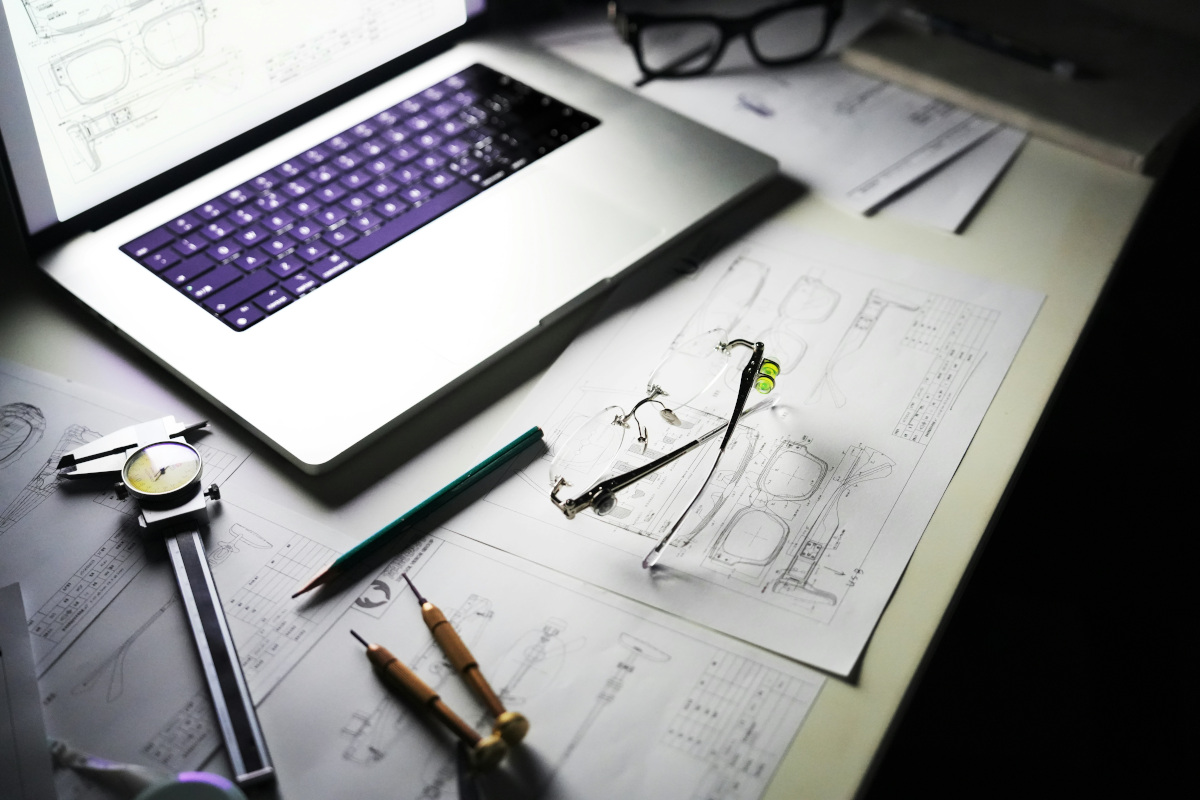 The image shows a desk setup with various items on it. In the center of the desk, there's an open laptop displaying what appears to be a blueprint drawing. Next to the laptop are several small models that resemble pairs of glasses, possibly used as reference for the design on the laptop screen. There's also a pair of measuring calipres laying closed, suggesting some recent activity. In front of the laptop is a pair of folded glasses, which might be used for comparison work or could be the photographer's own glasses. To the right of the laptop, there's an incomplete drawing or sketch on white paper, indicating that someone has been working on it. The desk itself is a simple, light-coloured surface, and the environment around it looks to be a workspace or home office.