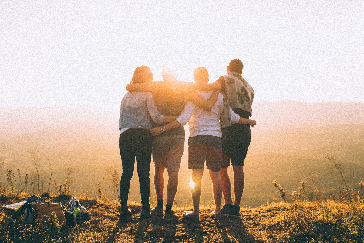 Four people stand in a line, their backs to the camera, with their arms wrapped around each other, looking at a sunset.