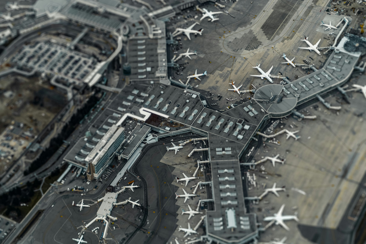 The image is an aerial photograph of an airport terminal. The terminal is characterized by multiple gates, each with its own assigned gate number and letter. Airplanes can be seen parked at their respective gates. The perspective of the image is from above, looking down upon the terminal and parking areas. A large expanse of the airport terminal is in view, with the foreground showing a bustling area where passengers might walk or wait for their flights. The ground appears to be a concrete surface with painted markings indicating parking spaces. The sky is visible but mostly obscured by buildings, suggesting that the airport is located near an urban area. There are no visible people or moving vehicles, giving the scene a somewhat still and calm appearance despite the scale of operations.