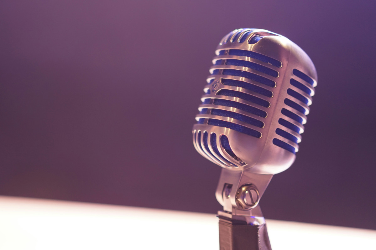The image shows a classic microphone on a stand, placed against a blue background. The microphone is silver and has a large diaphragm with the logo 'Shure' visible on it. It's positioned in front of what appears to be a black surface, possibly a podium or table. There are no texts overlaying the image, and the style seems to have a focus on the microphone, emphasizing its design and presence. The lighting is soft, casting gentle shadows behind the microphone, suggesting an indoor setting with artificial light.