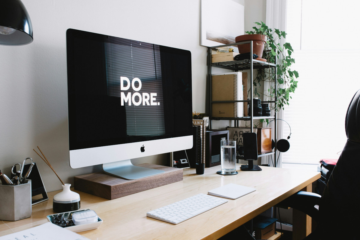 The image shows a modern home office setting. At the center is a large computer monitor displaying a motivational message, 'DO MORE.' in white letters on a black background. In front of the monitor is a wooden desk with a keyboard and mouse visible. On top of the desk, there are various items such as books, a glass of water, and some other objects that are not clearly identifiable. To the left of the desk, there's a white chair with a red cushion. The wall behind the monitor features a shelf holding more books and decorative items. To the right, there is a green plant adding a touch of nature to the space. The office appears well-organized and functional, suggesting a productive environment. The image has an overlay texture that resembles a piece of paper with a slight drop shadow effect, giving it a three-dimensional look.