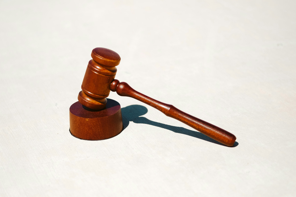 The image displays a wooden gavel and a wooden mallet against a neutral background. The gavel has a dark wood finish with a rectangular head and a flat, horizontal handle that appears to be made of the same wood material. It is placed slightly ahead and to the left of the mallet. The mallet also has a dark wood finish, but its surface is flat on both ends, with no distinct shape or design visible in this image. There are no texts or brands present in the image, and it has a realistic style typical for photographs meant to show an object clearly without any additional context