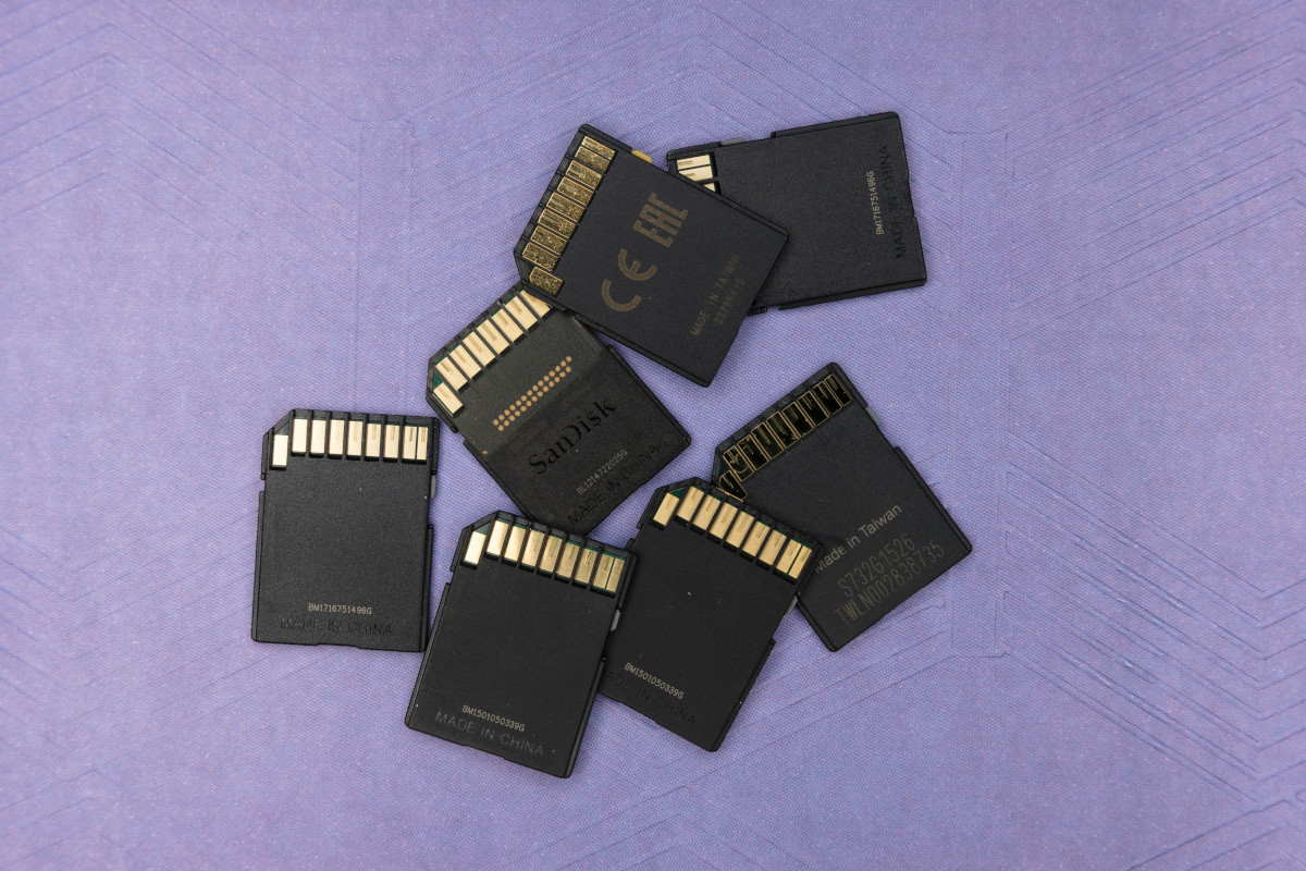 The image shows a collection of memory cards scattered in no particular order. These are small electronic devices used to store digital data, typically for cameras and other portable electronics. The cards are black in colour and have metallic gold contacts on their edges for connecting them to devices. The cards are scattered on a purple surface. There is no text providing additional context within the image itself.