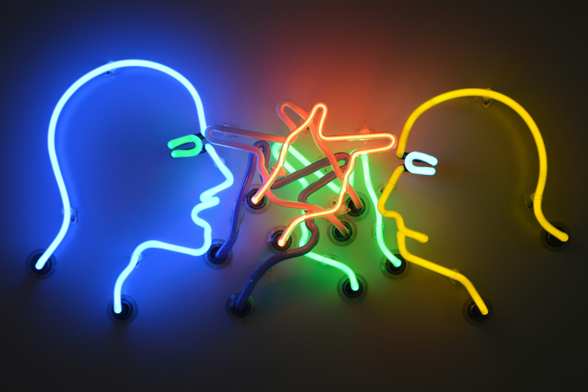 This is an image of a neon sign featuring two silhouettes of human heads. The sign has a playful or whimsical style, with one head being larger and positioned above the other, as if it's looking down at it. Each head is outlined in white, making them stand out against the blue background. Surrounding the silhouettes are various shapes and lines that appear to be drawn in neon pink and green colors, adding a touch of vibrancy to the sign. The overall impression is one of a fun, decorative piece that might be used to add character or theme to a bar, restaurant, or other establishment with a casual atmosphere.