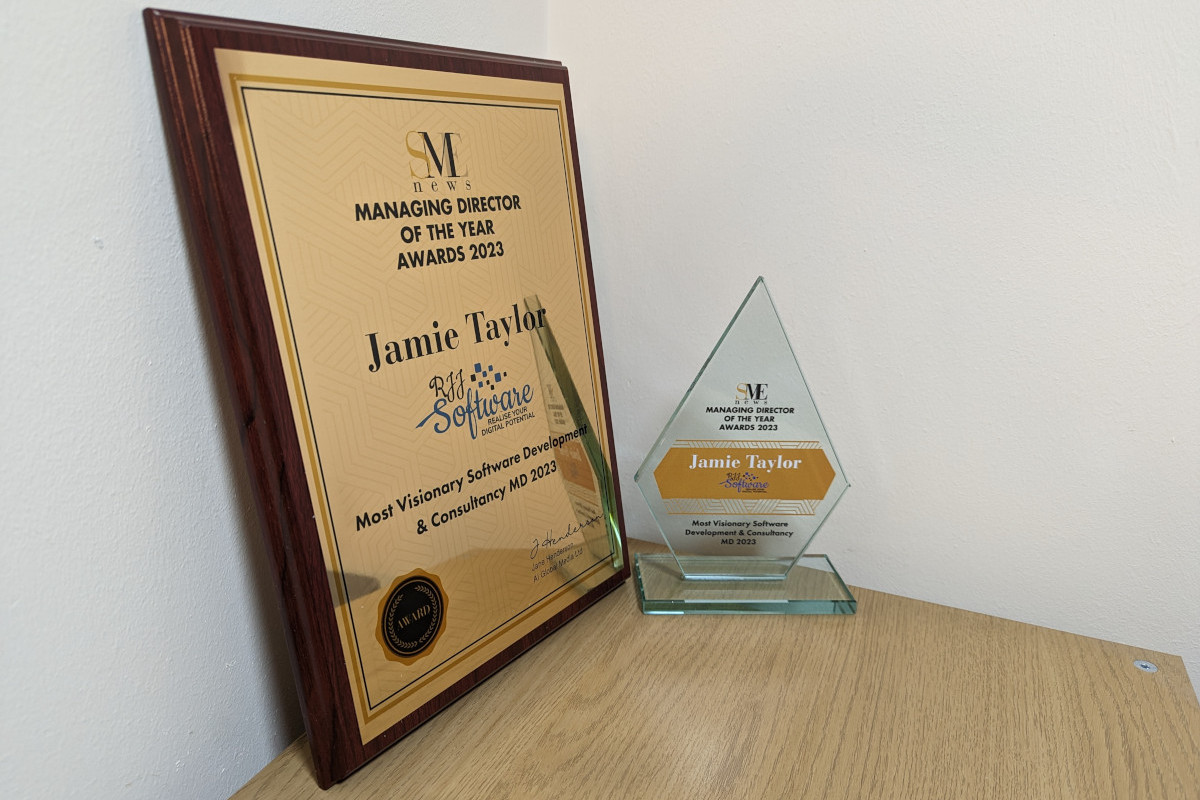 Both the award for Managing Director of the Year award for Most Visionary Software Development &amp; Consultancy MD and an award plaque. The award is made of a transparent glass, and is the shape of an elongated pentagon. It has the words &quot;SME News Managing Director of the year awards 2023&quot; in black text and all caps toward the top; Jamie's full name (jamie Taylor) and the RJJ Software logo on an orange/gold background in the centre; and the legend &quot;Most Visionary Software Development &amp; Consultancy MD 2023&quot; in black text and all caps at hte bottom. The wall plaque is a brass plate attached to a stained mahogany frame. It contains the same information that the award does.