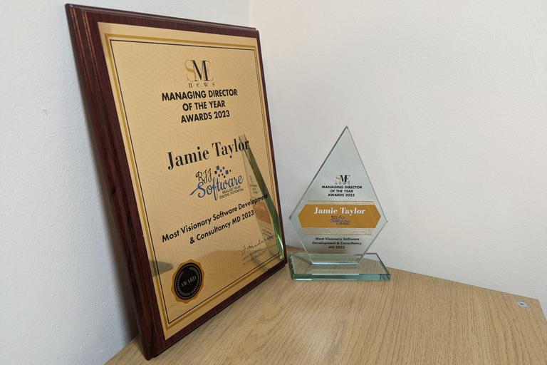 A close up of the award for Managing Director of the Year award for Most Visionary Software Development & Consultancy MD. The award is made of a transparent glass, and is the shape of an elongated pentagon. It has the words “SME News Managing Director of the year awards 2023” in black text and all caps toward the top; Jamie's full name (jamie Taylor) and the RJJ Software logo on an orange/gold background in the centre; and the legend “Most Visionary Software Development & Consultancy MD 2023” in black text and all caps at hte bottom.