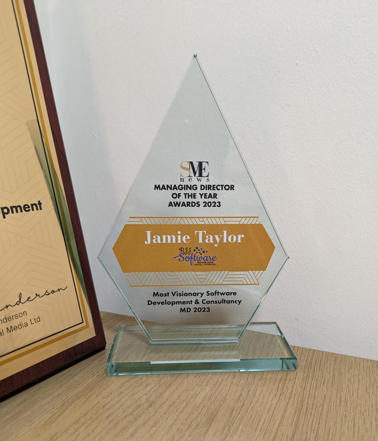 A close up of the award for Managing Director of the Year award for Most Visionary Software Development & Consultancy MD. The award is made of a transparent glass, and is the shape of an elongated pentagon. It has the words “SME News Managing Director of the year awards 2023” in black text and all caps toward the top; Jamie's full name (jamie Taylor) and the RJJ Software logo on an orange/gold background in the centre; and the legend “Most Visionary Software Development & Consultancy MD 2023” in black text and all caps at hte bottom.