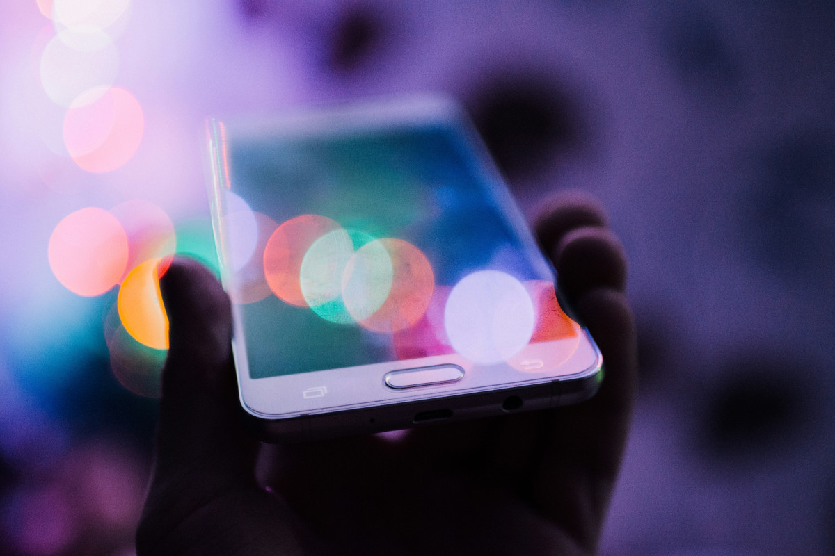 The image depicts a close-up view of a hand holding a smartphone with a colourful display. The screen is blurred, with various bright circles that resemble bokeh effect. In the background, out of focus, there are colourful lights that add to the artistic composition. A purple and black pattern overlay the image, giving it an abstract quality. The overall style of the image suggests a creative or artistic theme, possibly related to photography or visual art. 