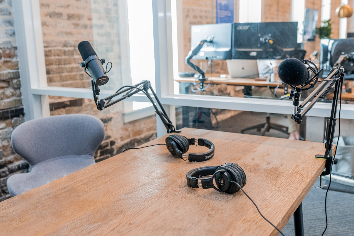 Two black headphones setup on brown wooden table, using microphone arms. Each microphone has a set of headphones plugged into them, and the microphones are arranged opposite of each other.