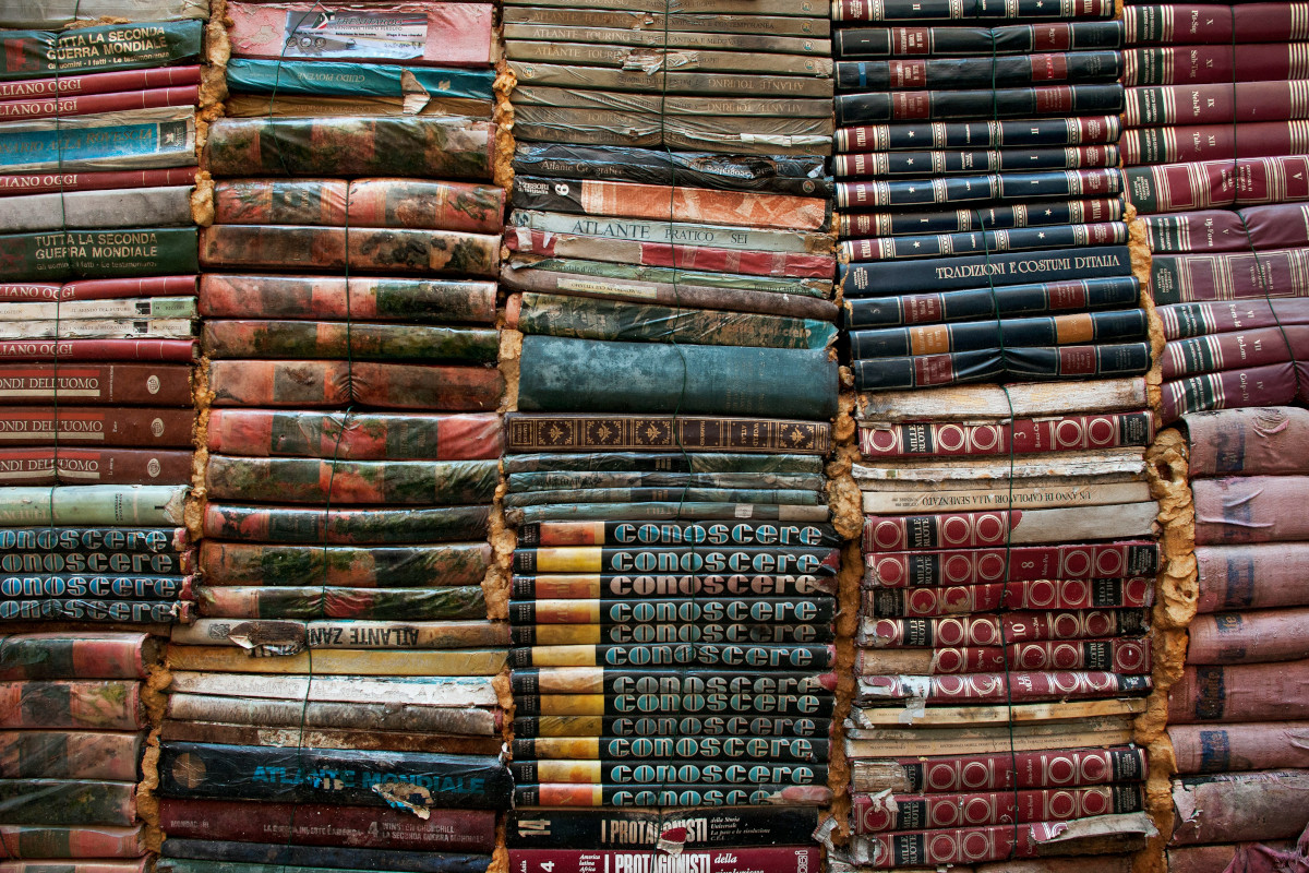 The image shows a collection of old books stacked on top of each other. There is a variety of book spines with visible titles and colours, which indicates a diverse range of subject matter or genres. Some spines are more prominently displayed than others due to the depth perspective as the viewer looks closer at the front of the stack. The books appear to be slightly worn, suggesting they may be vintage or well-used, and they have a rustic charm. The background is simple, with neutral tones that do not distract from the books. There are no people or distinguishable features in the background. The image has a naturalistic style and seems to capture a  moment of serendipity, perhaps in someone's personal library or a bookstore.