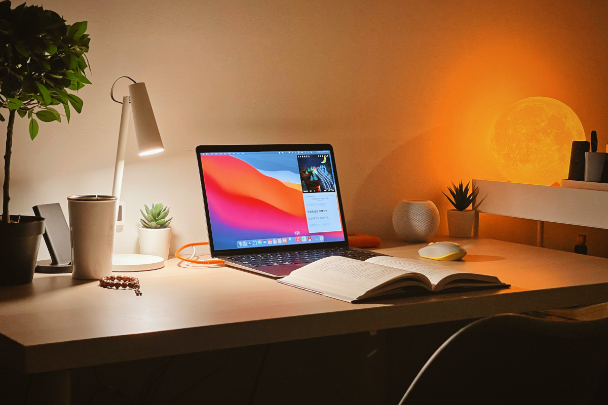 An open Macbook Pro sits on a white wooden desk. On the left is a lamp and some potted plants. On the right are more potted plants and a small shelf, behind the shelf is a lamp designed to look like the moon. In front of the Macbook Pro sits and open book. The entire image is taken at 3/4 profile, such that the desk and it's contents are not facing the camera.