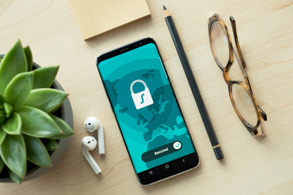 The image shows a smartphone displaying a digital interface with a padlock icon, suggesting that the phone is locked or protected. The screen also features a 'Sign In' button, which could imply that a user needs to enter their login credentials to unlock the phone. In front of the phone, on a wooden surface, there are objects suggesting a workspace: glasses, a potted plant (in the background), headphones with a cable hanging off the table, and a book. There is also a small amount of text visible at the bottom right corner of the phone screen that reads 'Secured.' The setting suggests someone's personal workstation, with a focus on privacy or security given the padlock image on the phone's screen.